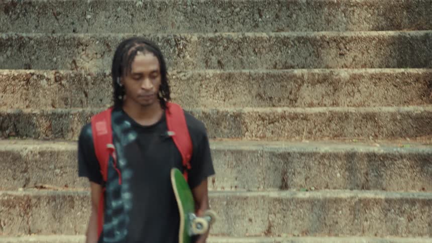 A young African American man with a skateboard and backpack walks down a flight of concrete stairs in slow motion