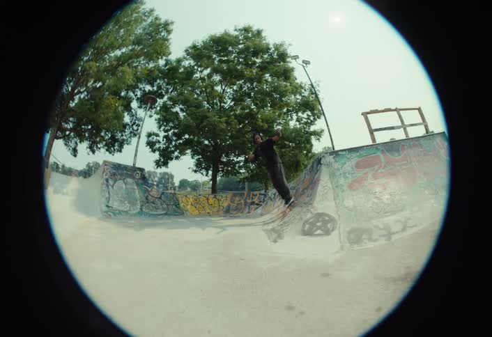 A slow-motion fisheye shot follows a man as he smoothly skates around a concrete bowl at an urban skatepark on a sunny day