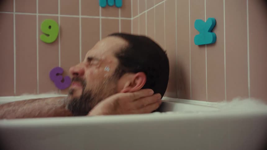 An exhausted man with a sticker on his face slowly sinks into a bubble bath, seeking a moment of peace in a child's bathroom