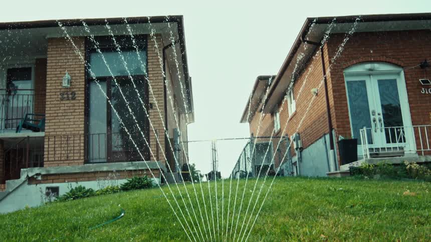 A low-angle slow-motion shot reveals a lawn sprinkler watering the front yard between two brick houses in a suburban neighborhood