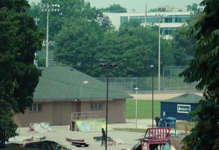 A wide tilt-down shot reveals a lone skateboarder practicing tricks in a public skatepark on a hazy day