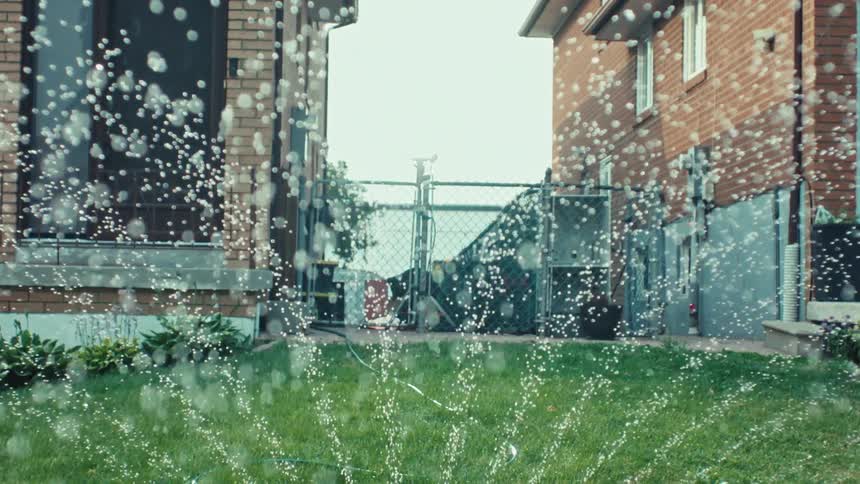A slow-motion close-up of a sprinkler watering a lush green lawn in a suburban backyard between two brick houses