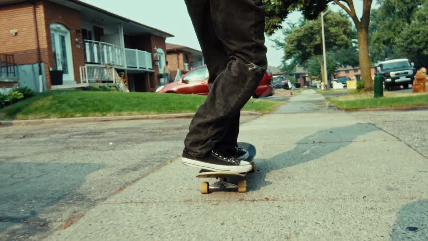 A low-angle, slow-motion shot follows a person's feet as they ride a skateboard down a suburban sidewalk