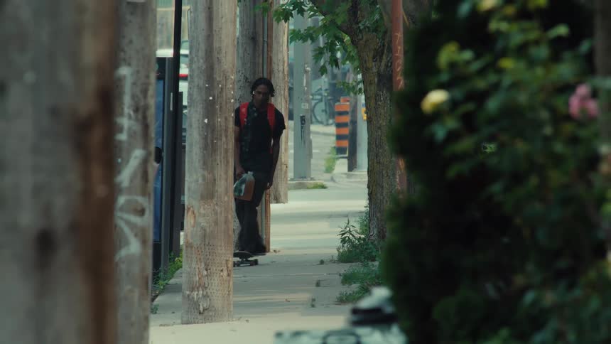 A person with dreadlocks and a backpack skateboards in slow motion down a city sidewalk lined with trees