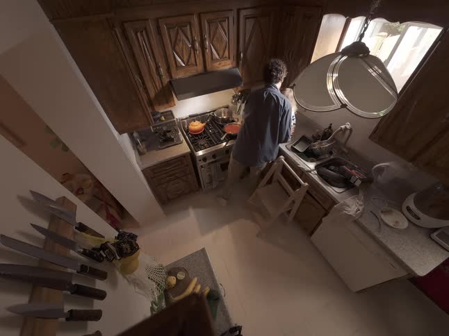 A father and his young daughter share a moment together in the kitchen while he cooks, filmed from a high wide-angle view