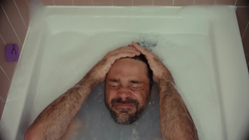 A distressed man fully clothed has an emotional breakdown in a bathtub, submerging himself in the water