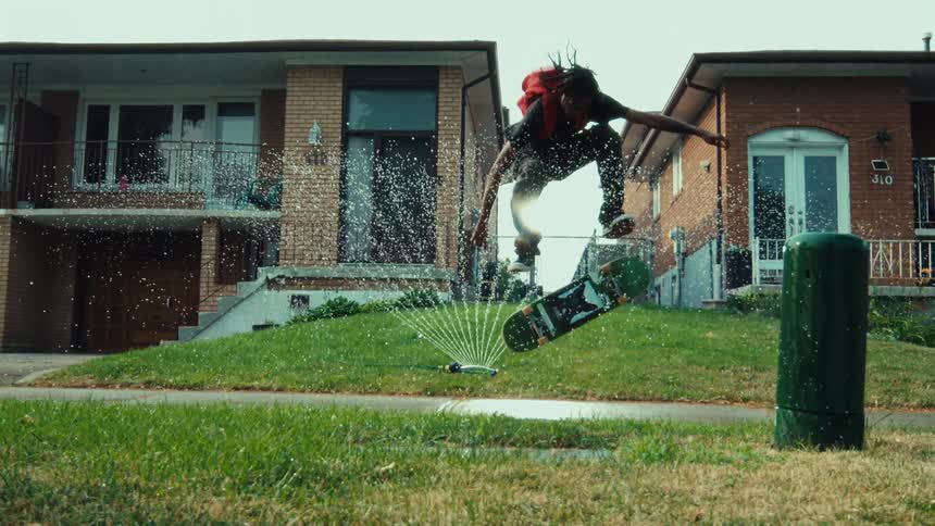 A skateboarder with a red backpack performs a kickflip over a lawn sprinkler in super slow motion on a summer day