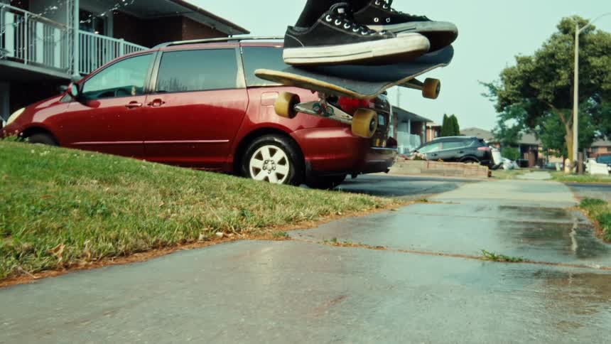 A skateboarder performs a kickflip in super slow motion on a wet suburban sidewalk as a lawn sprinkler sprays in the background