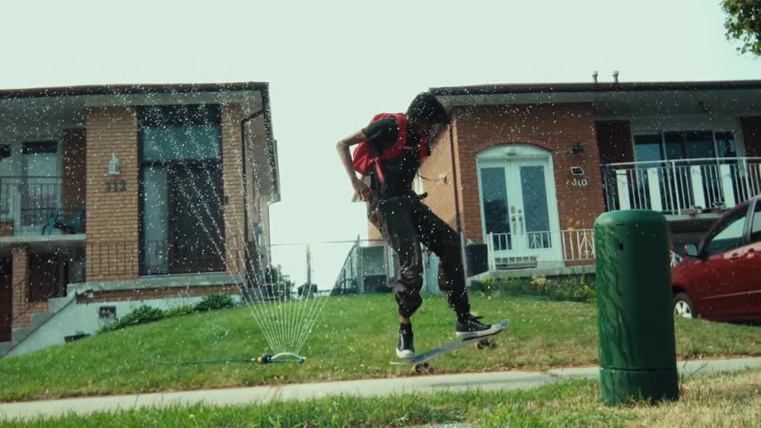 In super slow motion, a cool skateboarder with a red backpack jumps over a sprinkler while doing a kickflip trick on a suburban sidewalk
