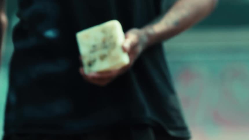 The camera tilts up from a blurry skateboard to a skater holding a block of wax with the explicit phrase 'Fuck Yourself' written on it