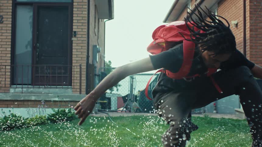A carefree young man with dreadlocks and a red backpack playfully dances in a lawn sprinkler in slow motion