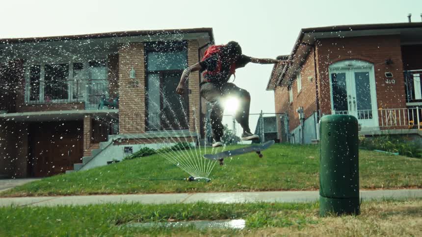 A skateboarder performs a kickflip in super slow motion, soaring through the mist of a lawn sprinkler on a sunny suburban street