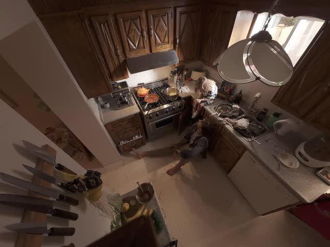 An exhausted man sits on the floor while a woman sits on the counter in their messy kitchen as dinner cooks on the stove