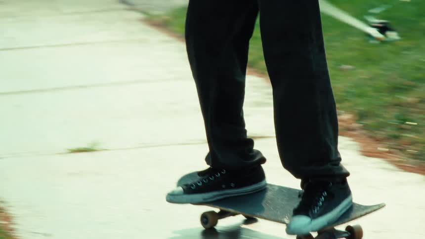 In slow motion, a skateboarder does a kickflip trick while riding through a lawn sprinkler on a sidewalk