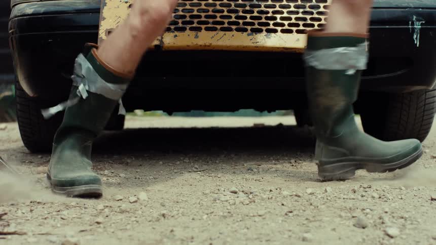 A low-angle shot of a person's legs in duct-taped rubber boots as they frantically run away from a car