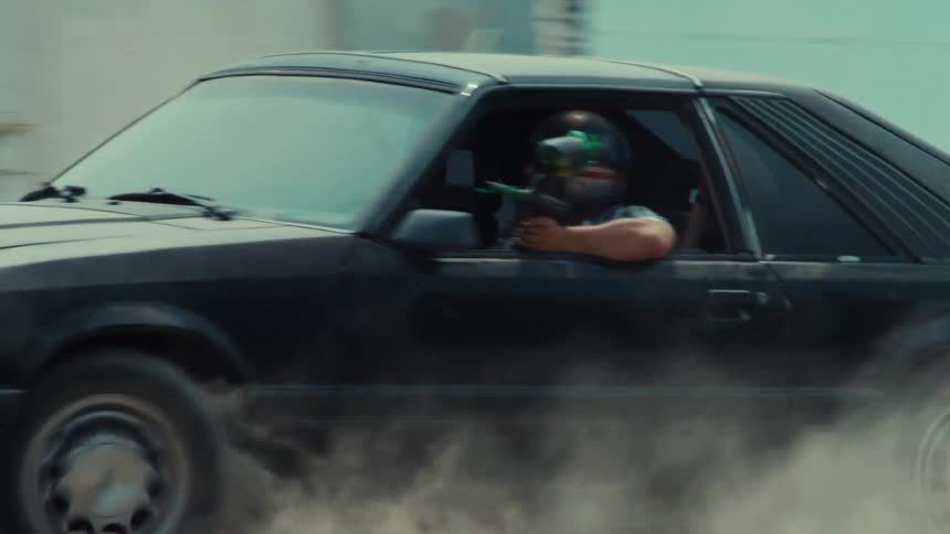 A man aims a paintball gun from a speeding black muscle car as it kicks up dust on a dirt road