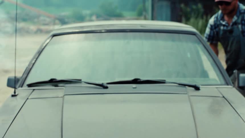 A serious-looking farmer wearing overalls gets into his old, dusty car on a farm