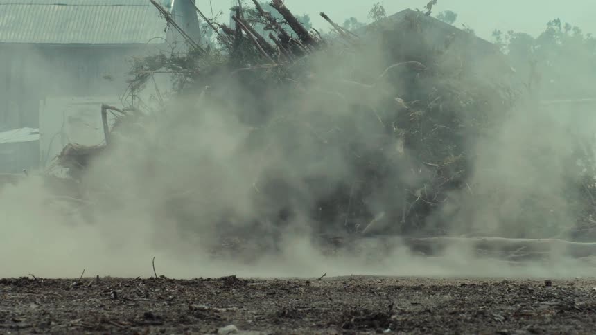 Two cars race down a dirt road in slow motion, kicking up a massive cloud of dust in a rural junkyard
