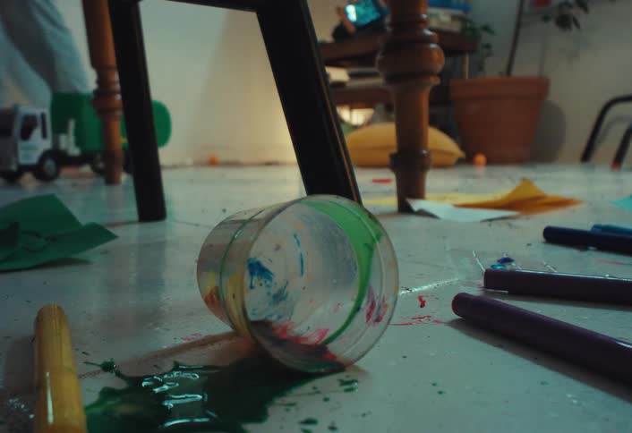 A cup of green paint is spilled on the floor in a messy room filled with art supplies and toys, while a person walks in the background