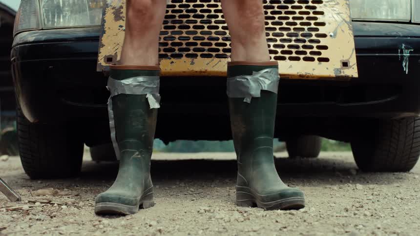 A low angle shot of a person's legs in green rubber boots secured with duct tape, standing in front of an old car