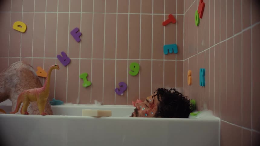 A bloodied man with stickers on his face lies unresponsive in a bubble bath surrounded by colorful alphabet magnets and toys