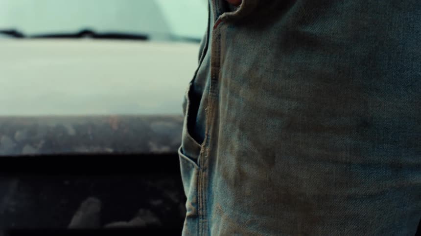 A close-up shot shows a man's hand slowly putting a set of keys into the pocket of his dirty denim jeans