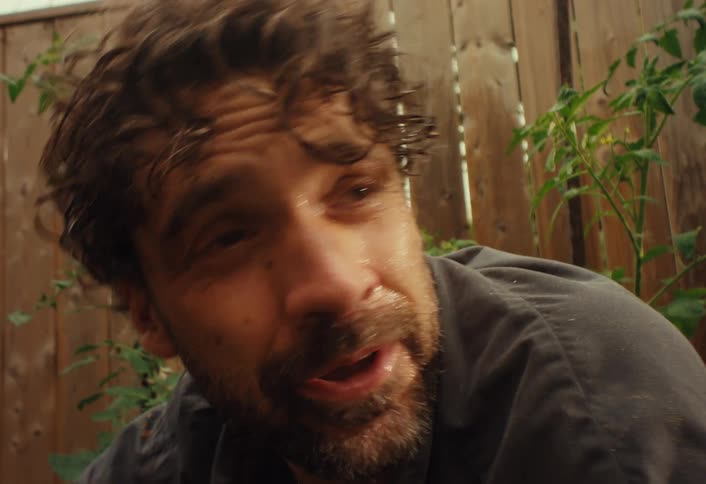A sweaty and distraught man, covered in dirt, has an emotional breakdown in a garden, wiping his tears with a dirty hand