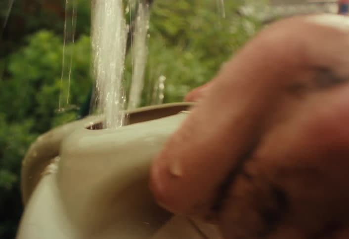 A gardener with dirt on their hands fills a watering can using a yellow hose in a lush backyard garden