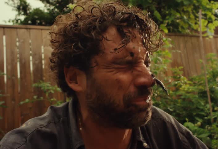 A man with a dirty face laughs in slow motion as a child's hand smears more mud on him while they play in a backyard garden