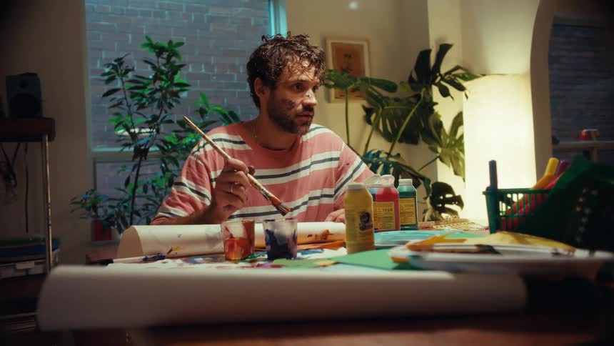 A bearded artist with paint on his face sits at a messy table and dips his brush into paint while working on a project at home