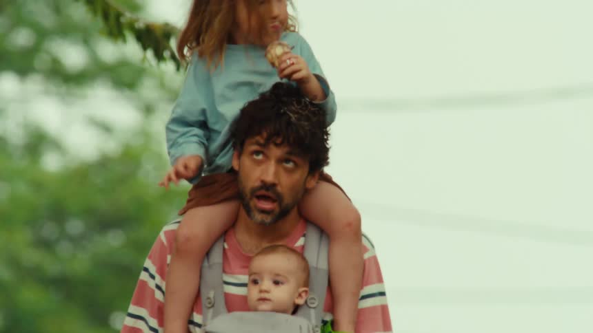 An exhausted father walks with his baby in a carrier and his toddler eating an ice cream cone on his shoulders