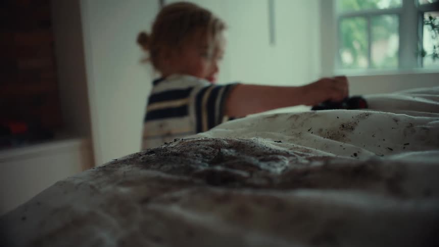 A mischievous toddler plays with a toy monster truck in dirt on white bedsheets in a bedroom