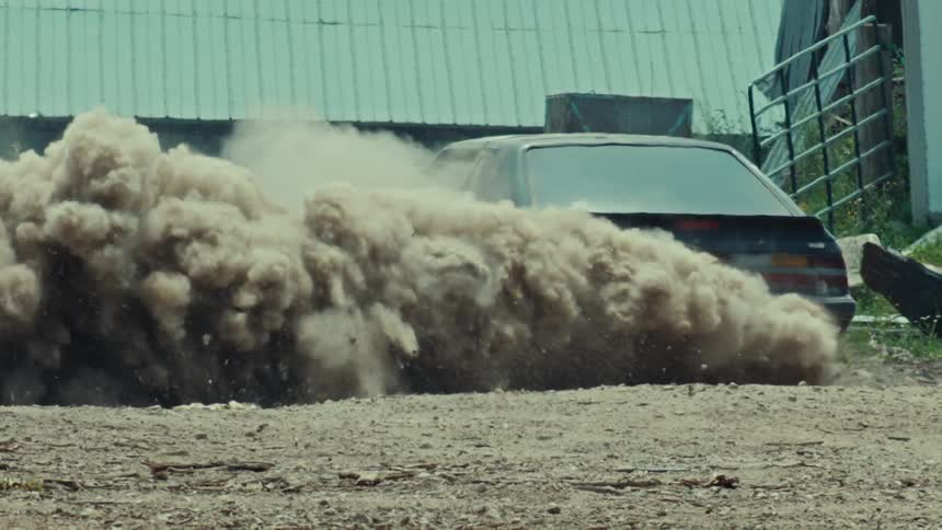A black muscle car performs a burnout on a dirt road, kicking up a massive cloud of dust in slow motion