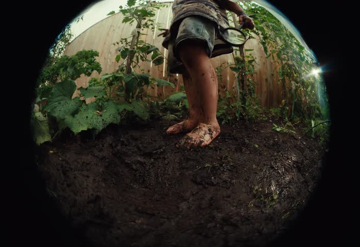 A curious toddler covered in mud stomps barefoot in a backyard garden in a slow-motion fisheye shot
