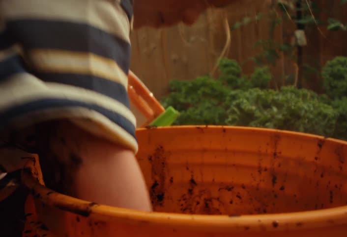 A close-up slow motion shot shows a young boy having fun getting messy while playing with a bucket full of mud in the backyard garden