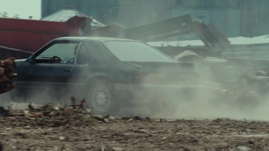 A classic black muscle car drives and drifts in slow motion on a dirt road, kicking up a large cloud of dust