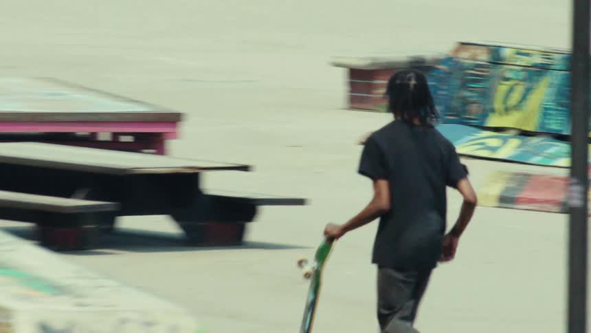 A young man with dreadlocks effortlessly performs a kickflip trick on his skateboard at a DIY urban skatepark in slow motion