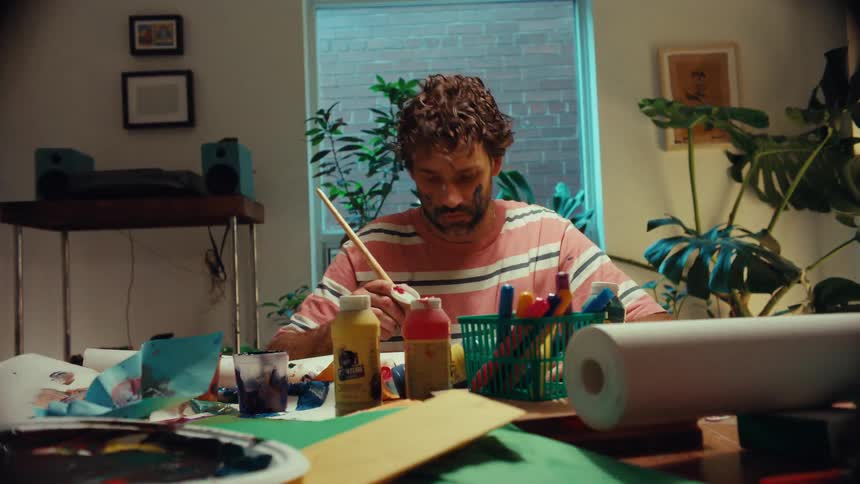 A focused artist with paint on his face works at a messy table in his studio, while a colorful pinwheel spins in the foreground