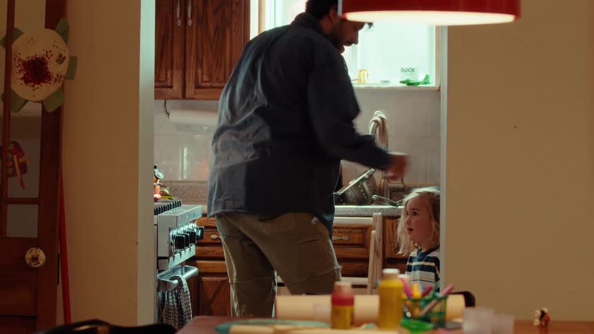 A father and his young daughter share a playful moment in their warm, sunlit kitchen while he washes dishes