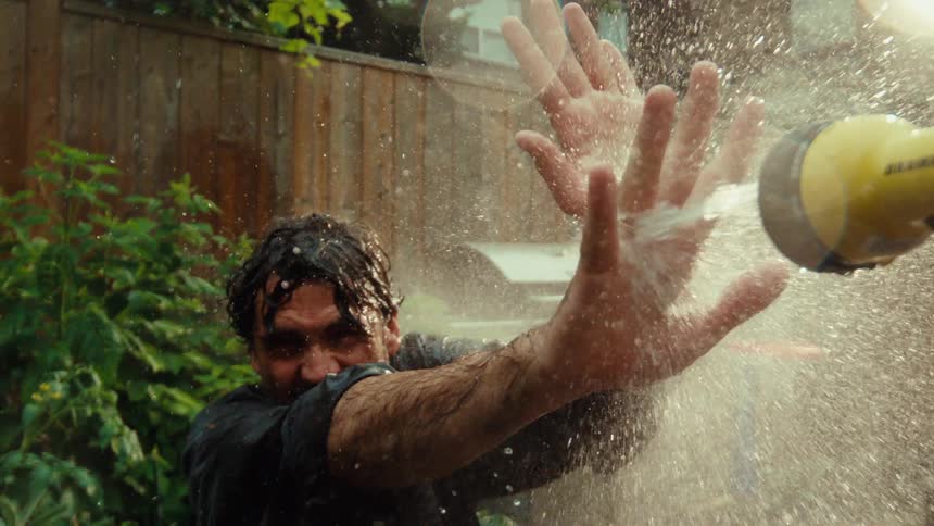 Playful man gets drenched by a garden hose held by his child during a fun summer water fight in the backyard, captured in cinematic slow motion
