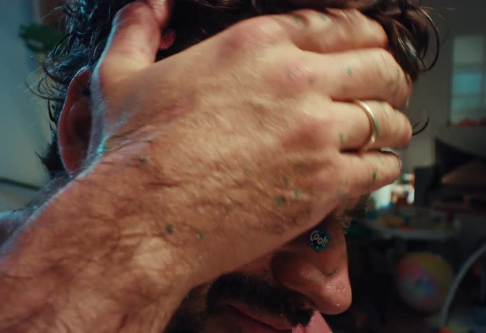 An exhausted father, covered in children's stickers and paint, rubs his face in distress in a messy room