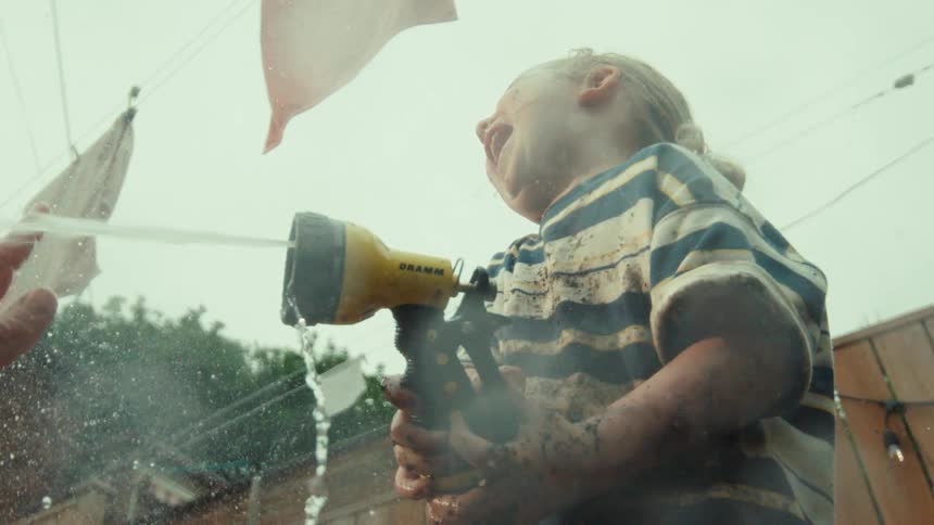 From a low angle perspective, a gleeful toddler covered in mud laughs while spraying a garden hose in slow motion