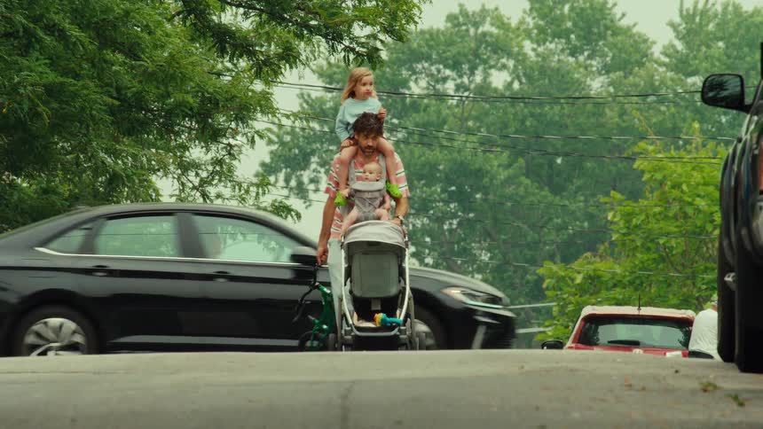 A super dad walks down a residential street carrying a toddler on his shoulders and a baby in a carrier, while also pushing a stroller and carrying a bike