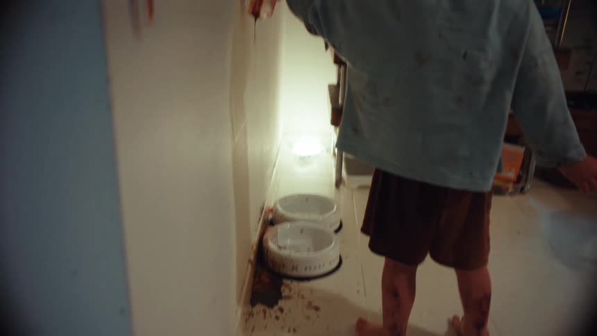 A mischievous barefoot girl covered in dirt creates a mess by dripping dark paint from a jar down a white wall in her home