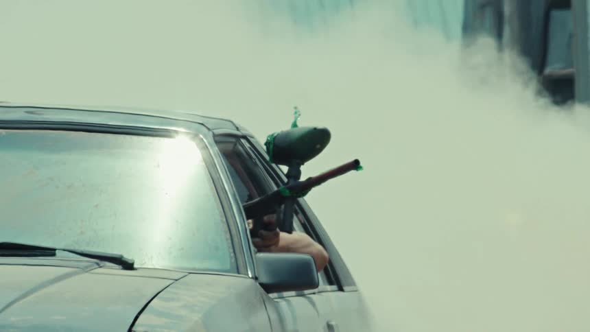 A paintball player aims their marker out of a car window while engulfed in a cloud of smoke in dramatic slow motion