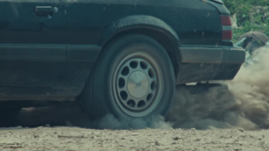 A close-up slow-motion shot of a black vintage car's tire spinning and doing a burnout, kicking up a massive cloud of dust from a dirt road