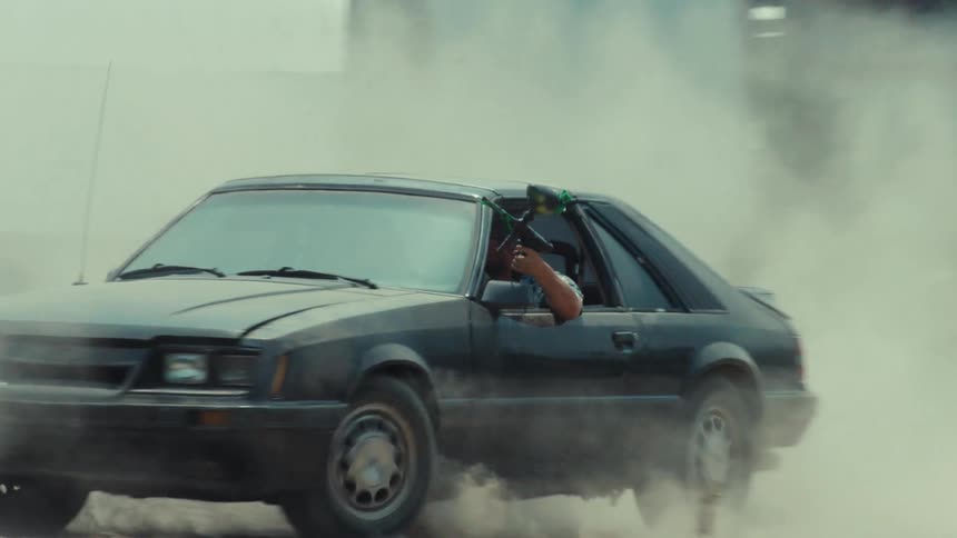 Two cars engage in a high-speed chase through a dusty industrial lot while a man shoots a paintball gun from a vintage sports car in slow motion