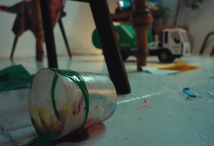 A close-up shot of a spilled cup of green paint on a messy floor with children's toys as the camera racks focus to the background