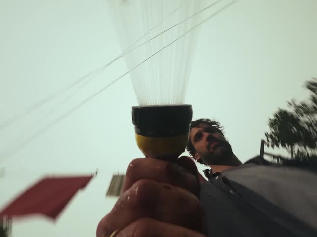 Low-angle point-of-view shot of a man holding a garden hose nozzle as it sprays water into the air in a backyard