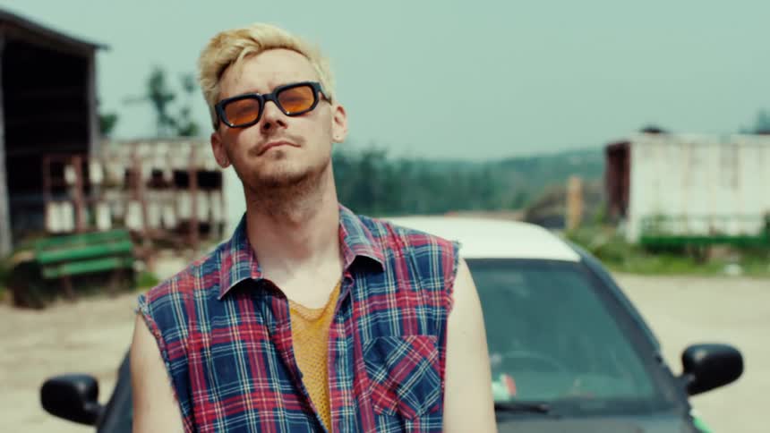 A confident young man with bleached blonde hair and sunglasses poses in front of a car in a rural setting, then walks out of frame